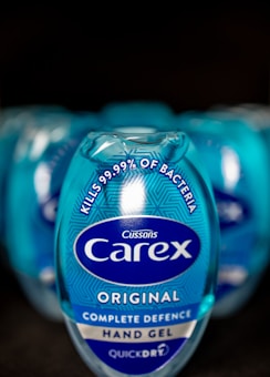 A close-up view of a blue Carex hand gel bottle prominently displaying text that states it kills 99.99% of bacteria. The bottle design includes the Carex logo and branding elements on a shiny, reflective surface. Multiple bottles are blurred in the background, suggesting a commercial arrangement.
