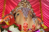 A detailed sculpture of a deity adorned with intricate jeweled and pearl decorations. Surrounding the statue are vibrant flowers and ribbons, set against a rich pink and gold drapery background.