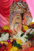 A vividly decorated statue of Lord Ganesha, adorned with intricate jewelry and vibrant floral garlands. The statue has detailed ornamentation on the headpiece, with gold elements and embedded pearls, set against a backdrop of rich pink drapery.
