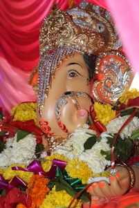 A vividly decorated statue of Lord Ganesha, adorned with intricate jewelry and vibrant floral garlands. The statue has detailed ornamentation on the headpiece, with gold elements and embedded pearls, set against a backdrop of rich pink drapery.