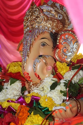 A vividly decorated statue of Lord Ganesha, adorned with intricate jewelry and vibrant floral garlands. The statue has detailed ornamentation on the headpiece, with gold elements and embedded pearls, set against a backdrop of rich pink drapery.