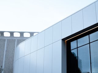 An architectural shot showcasing a modern building with clean, sleek lines and large glass windows. The structure features a combination of white panels and reflective glass surfaces, complemented by a portion of a neighboring building with a unique perforated design.