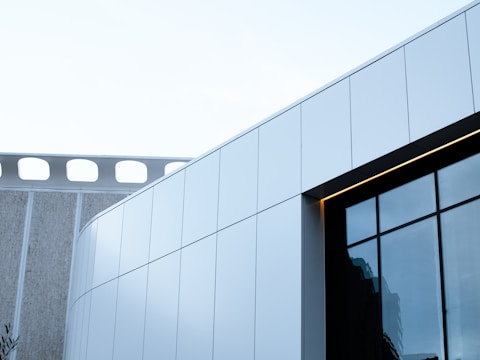 An architectural shot showcasing a modern building with clean, sleek lines and large glass windows. The structure features a combination of white panels and reflective glass surfaces, complemented by a portion of a neighboring building with a unique perforated design.