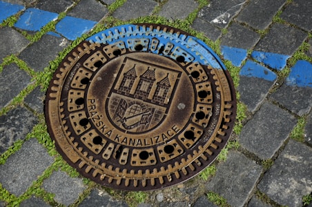The image features a metal manhole cover on a cobblestone street with patches of green grass growing between the stones. The manhole cover is engraved with architectural motifs and the text 'PRAŽSKA KANALIZACE' around its perimeter. A blue painted line intersects the cobblestones, partially covering the manhole cover.