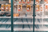 A refrigerated display case in a store with mostly empty shelves, except for a few pre-packaged meat products hanging and some items positioned on lower shelves. The focus is on the scarcity of products available.