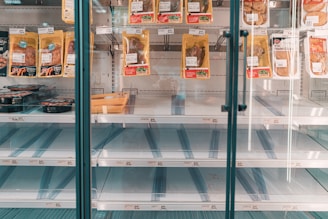 A refrigerated display case in a store with mostly empty shelves, except for a few pre-packaged meat products hanging and some items positioned on lower shelves. The focus is on the scarcity of products available.