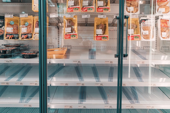 A refrigerated display case in a store with mostly empty shelves, except for a few pre-packaged meat products hanging and some items positioned on lower shelves. The focus is on the scarcity of products available.