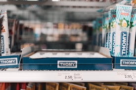 A store shelf with multiple tubes of Thomy mayonnaise displayed in a cardboard holder. The tubes are predominantly white with blue and green accents, featuring images of eggs and herbs. Price tags are visible on the shelf below, indicating the cost of the items.