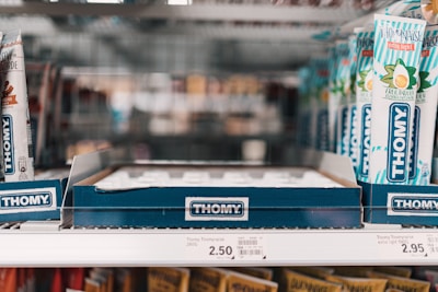 A store shelf with multiple tubes of Thomy mayonnaise displayed in a cardboard holder. The tubes are predominantly white with blue and green accents, featuring images of eggs and herbs. Price tags are visible on the shelf below, indicating the cost of the items.