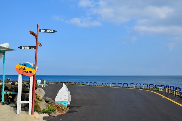 A coastal road curves gently with blue ocean water in the background. Multiple signposts are visible, with one pointing towards Japan and others showing local destinations in Korean and English. A colorful sign reads 'Jeju Halryang.' The sky is partly cloudy, and there are guardrails along the road.