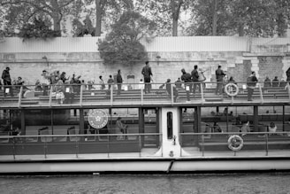 A riverboat filled with passengers cruises along a waterway. People are seated and standing on the upper deck, observing the surroundings. The boat is near a stone embankment, with trees and a wall in the background.