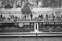 A riverboat filled with passengers cruises along a waterway. People are seated and standing on the upper deck, observing the surroundings. The boat is near a stone embankment, with trees and a wall in the background.