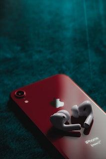 A pair of wireless earbuds rests on the back of a red smartphone, which features the Apple logo. The phone is placed on a textured dark green surface, creating a striking contrast between the bright red device and the deep background.