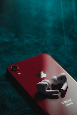 A pair of wireless earbuds rests on the back of a red smartphone, which features the Apple logo. The phone is placed on a textured dark green surface, creating a striking contrast between the bright red device and the deep background.