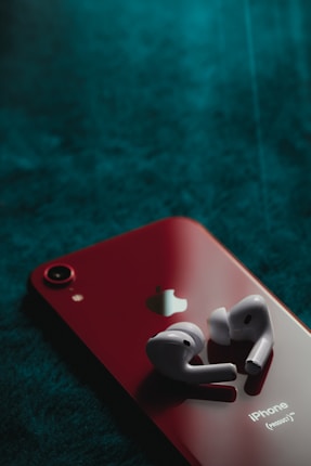 A pair of wireless earbuds rests on the back of a red smartphone, which features the Apple logo. The phone is placed on a textured dark green surface, creating a striking contrast between the bright red device and the deep background.