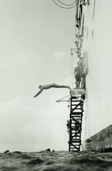 A person is diving off a platform attached to the side of a large vessel. The platform consists of metal steps, and several other people are seen preparing to dive. The sea is calm, and the angle of the photo captures the diver mid-air above the water.