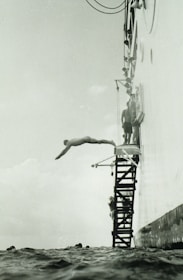 A person is diving off a platform attached to the side of a large vessel. The platform consists of metal steps, and several other people are seen preparing to dive. The sea is calm, and the angle of the photo captures the diver mid-air above the water.
