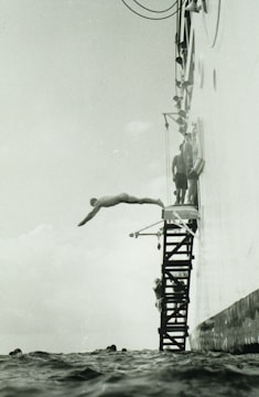 A person is diving off a platform attached to the side of a large vessel. The platform consists of metal steps, and several other people are seen preparing to dive. The sea is calm, and the angle of the photo captures the diver mid-air above the water.