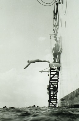 A person is diving off a platform attached to the side of a large vessel. The platform consists of metal steps, and several other people are seen preparing to dive. The sea is calm, and the angle of the photo captures the diver mid-air above the water.
