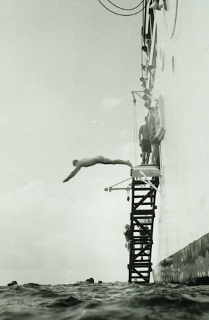 A person is diving off a platform attached to the side of a large vessel. The platform consists of metal steps, and several other people are seen preparing to dive. The sea is calm, and the angle of the photo captures the diver mid-air above the water.