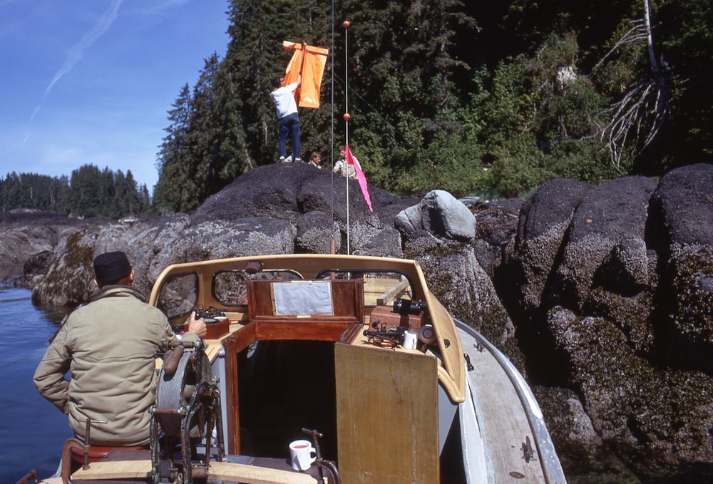 Mann in brauner Jacke und brauner Hose steht neben einem braunen Holzboot auf einem felsigen Hügel während