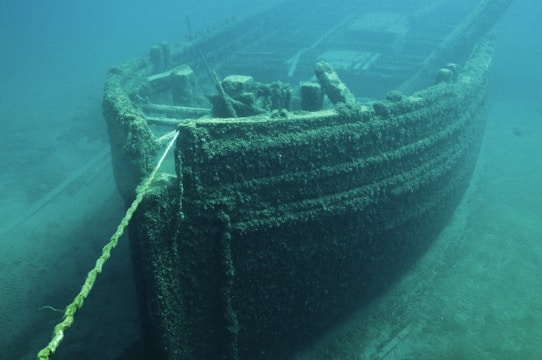 A sunken ship partially covered in algae and marine growth is visible underwater. The vessel's front is prominently shown, with a rope extending from it. The water around is a clear blue-green, allowing the details of the ship's structure and the sandy seabed to be seen.