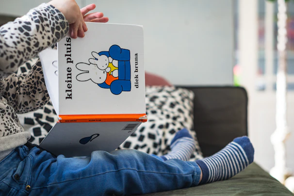 A child is sitting on a sofa, holding and presumably reading a children's book titled 'kleine pluis' by Dick Bruna. The child is wearing striped socks, blue jeans, and a sweater with a black and white pattern. The book features a cartoon of a rabbit on its cover. The background includes a cushion with a black and white leopard print design.