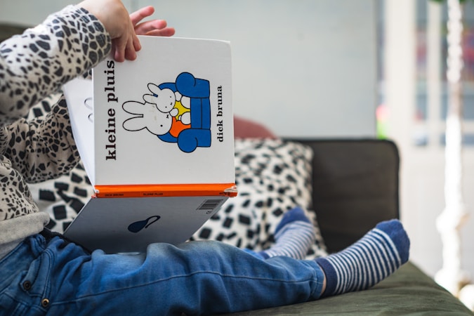 A child is sitting on a sofa, holding and presumably reading a children's book titled 'kleine pluis' by Dick Bruna. The child is wearing striped socks, blue jeans, and a sweater with a black and white pattern. The book features a cartoon of a rabbit on its cover. The background includes a cushion with a black and white leopard print design.