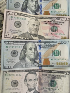 A collection of US dollar bills of various denominations, including $5, $50, and $100 notes. The bills are neatly arranged with visible portraits of US historical figures: Abraham Lincoln on the $5 bill, Ulysses S. Grant on the $50 bill, and Benjamin Franklin on the $100 bill. The design elements and colors of the bills are clearly visible, with security features like watermarks and color-changing ink.