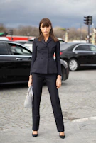 Woman confidently walking down a cobblestone street in a sleek black suit with a flowing cream blouse underneath.