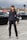 A woman is standing on a cobblestone street wearing a tailored black suit with a double-breasted jacket and matching trousers. She holds a gray handbag with gold accents and wears black pointed-toe heels. The background features blurred traffic lights and several black cars, with an overcast sky above.