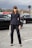 A woman is standing on a cobblestone street wearing a tailored black suit with a double-breasted jacket and matching trousers. She holds a gray handbag with gold accents and wears black pointed-toe heels. The background features blurred traffic lights and several black cars, with an overcast sky above.