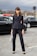 A woman is standing on a cobblestone street wearing a tailored black suit with a double-breasted jacket and matching trousers. She holds a gray handbag with gold accents and wears black pointed-toe heels. The background features blurred traffic lights and several black cars, with an overcast sky above.