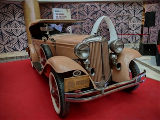 A vintage car with a classic design is prominently displayed on a red carpet. The car features a beautiful grille, large round headlights, and a pale beige color. The background includes a patterned wall and a sign promoting a selfie opportunity with the vintage car.