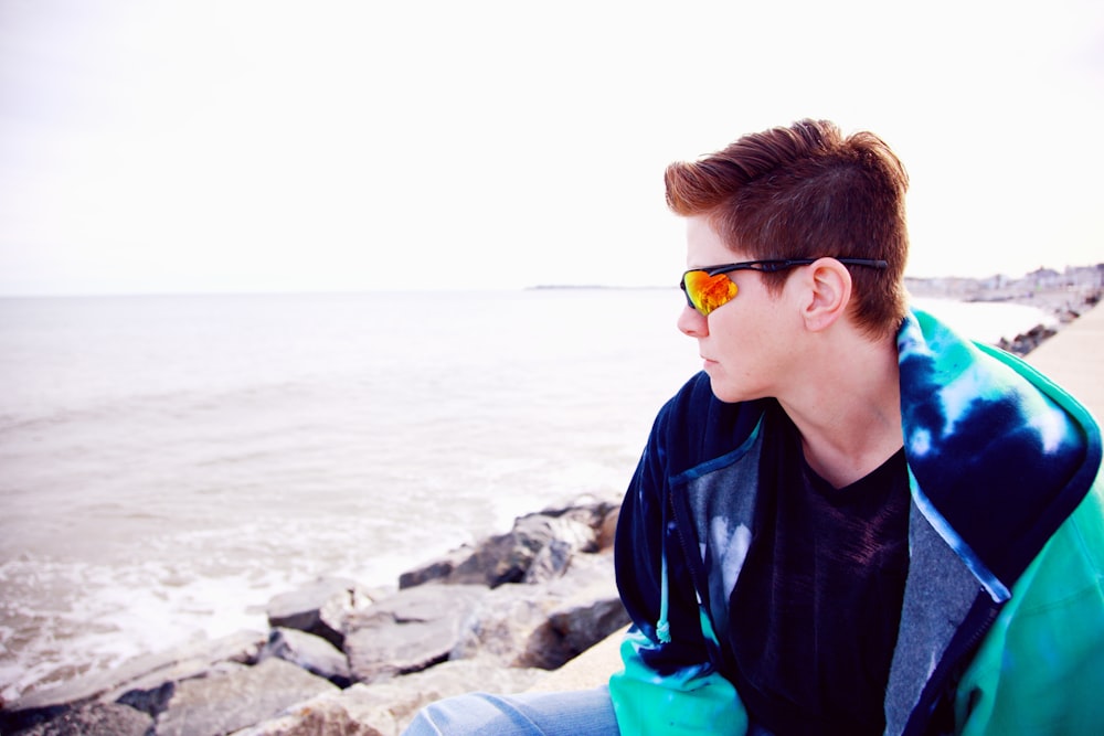 낮 동안 바다 근처 바위에 앉아 선글라스를 끼고 있는 파란색 재킷을 입은 소년