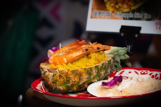 A dish of pineapple fried rice is served in a halved pineapple, topped with large shrimp. The rice is vibrant yellow and garnished with purple flower petals. Next to it, there is a plate with more rice and flower embellishments. The presentation is colorful and appetizing.