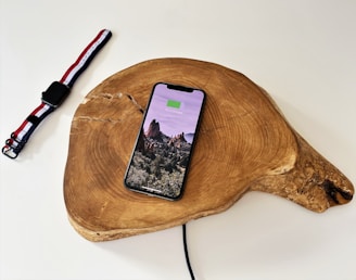 A smartphone displaying a landscape image and battery charging status rests on a wooden charging pad. Nearby, a smartwatch with a red, white, and black striped band lies on a white surface. A charging cable is visible connecting to the wooden pad.