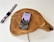 A smartphone displaying a landscape image and battery charging status rests on a wooden charging pad. Nearby, a smartwatch with a red, white, and black striped band lies on a white surface. A charging cable is visible connecting to the wooden pad.
