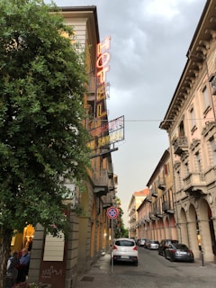 A narrow street flanked by classic European-style buildings with balconies. A neon 'HOTEL' sign is prominently displayed on the side of a building on the left. Cars are parked along the cobblestone street, and there is a no parking sign visible. The sky is overcast, and a tree partially obscures the building with the hotel sign.