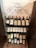 A black wrought iron wine rack holds several bottles of wine arranged neatly. The bottles have labels with various designs and are sealed with red or orange caps. The rack is placed against a light-colored wall, and the floor is covered with terracotta tiles.