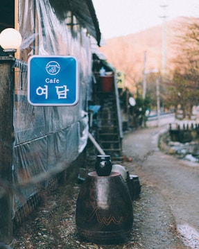 A rustic outdoor pathway with a small caf&eacute; sign in Korean and English. The setting includes wooden structures and a large decorative jar in the foreground. The path is lined with plants and appears rural and peaceful.