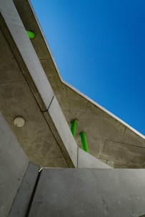 A modern architectural structure composed of concrete and featuring angular lines. Green cylindrical elements contrast with the concrete, while a bright blue sky provides a backdrop, highlighting the geometric design.
