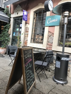 An outdoor caf&eacute; setting with a rustic ambiance. The scene features a blackboard menu listing daily soups and desserts, placed on a cobblestone pathway. Two folding chairs with cushions are positioned around a small table. Adjacent stands a tall outdoor heater. The caf&eacute;'s exterior is composed of weathered stone and brick, with a window displaying bread loaves inside. There are potted plants adding greenery to the scene.