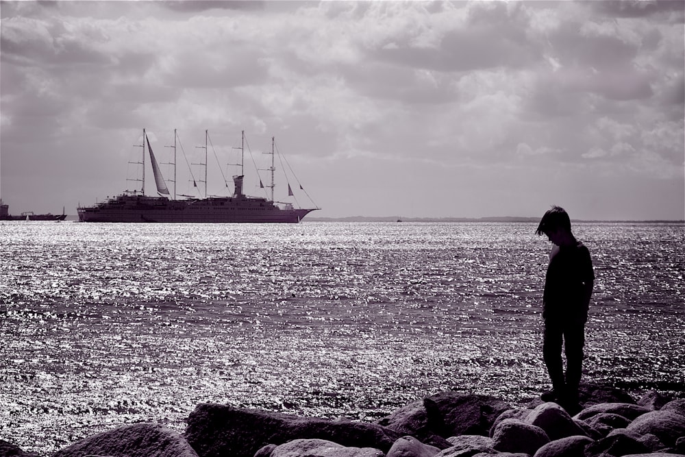 grayscale photo of man standing on rock near body of water