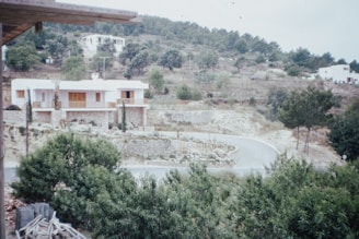 A scenic hillside area with a curving road leading up to white modern houses integrated into a lush green landscape. The vegetation is abundant with trees and shrubs, alongside stone terraces. The setting conveys a peaceful rural or suburban environment with a touch of architectural elegance.