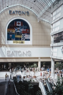 A shopping mall interior with a large Eaton's sign prominently displayed. Flags are hung in an arch above the sign. People are walking around and shopping, with planters and escalators visible in the space. The ceiling is made of glass, allowing natural light to illuminate the area.