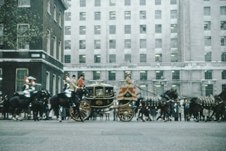 A procession featuring a horse-drawn carriage escorted by soldiers riding horses. The carriage is ornate, with gold decorations, and is flanked by horses adorned in ceremonial gear. Behind the procession, a large, historic building with several windows stands prominently. The scene appears to take place on a city street with some onlookers in the background.