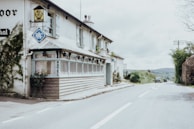 A countryside motel is situated along a quiet rural road. The building features traditional architecture with white walls and a wooden porch. Nearby, there are signs for the AA and RAC, indicating the presence of a hotel. The road is lined with greenery, and there are a few vintage cars parked in the distance.
