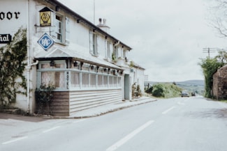 A countryside motel is situated along a quiet rural road. The building features traditional architecture with white walls and a wooden porch. Nearby, there are signs for the AA and RAC, indicating the presence of a hotel. The road is lined with greenery, and there are a few vintage cars parked in the distance.