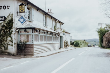 A countryside motel is situated along a quiet rural road. The building features traditional architecture with white walls and a wooden porch. Nearby, there are signs for the AA and RAC, indicating the presence of a hotel. The road is lined with greenery, and there are a few vintage cars parked in the distance.
